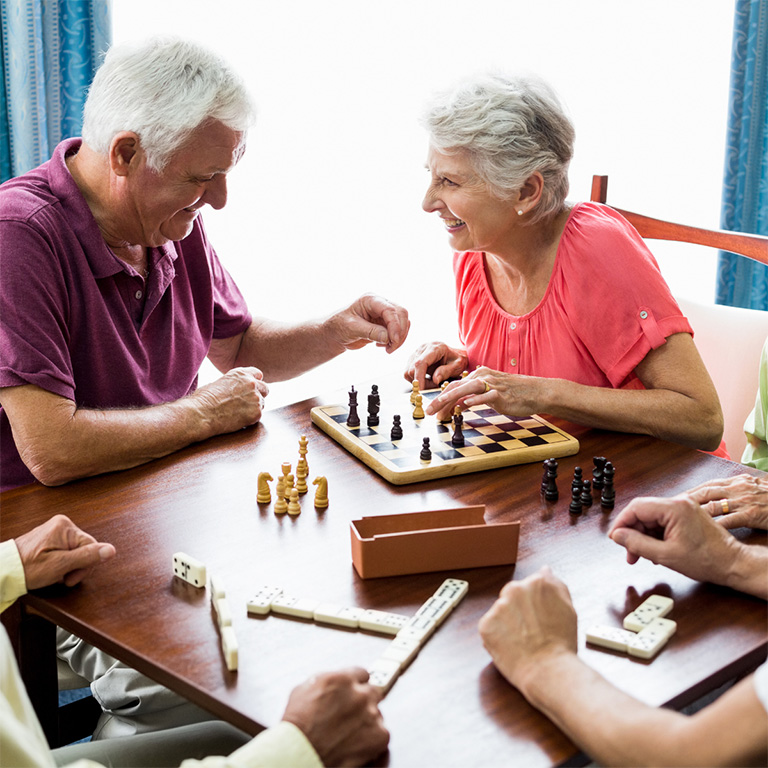 Group of smiling seniors playing board games together at a table, including chess and dominoes, enjoying social time in a bright room