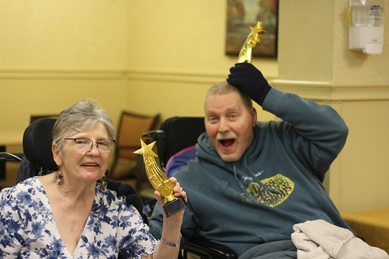 An elder woman and a man, both seated in wheelchairs, are smiling and holding up gold star-shaped trophies.