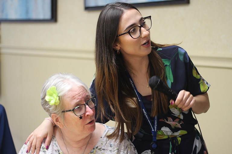 A younger woman with long brown hair and glasses is holding a microphone and standing beside an older woman with white hair, glasses, and a green flower clip. The younger woman has her arm around the older woman, who has a star sticker on her cheek.