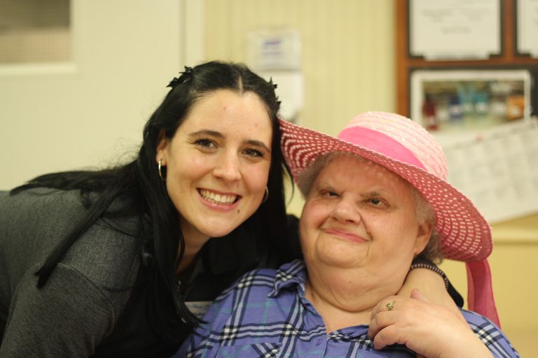 A smiling younger woman with long dark hair and a grey top is hugging an older woman with white hair wearing a pink sunhat and a blue plaid shirt.