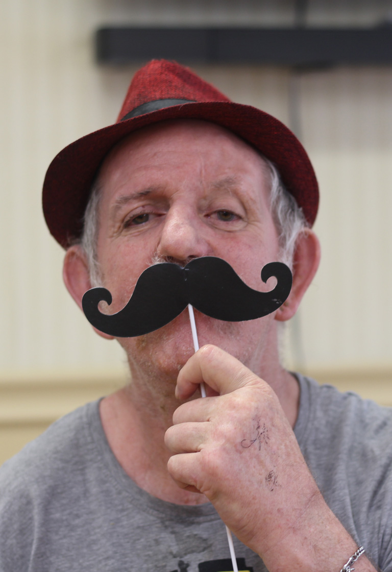 An elder man wearing a red hat and grey T-shirt is playfully holding a paper moustache on a stick in front of his face. He is smiling slightly and posing indoors in a light-colored room.