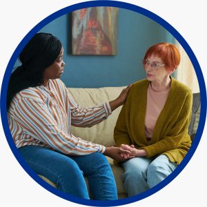 A personal support worker sits on a couch beside an older woman, gently holding her hand and resting a hand on her shoulder, offering comfort and support.